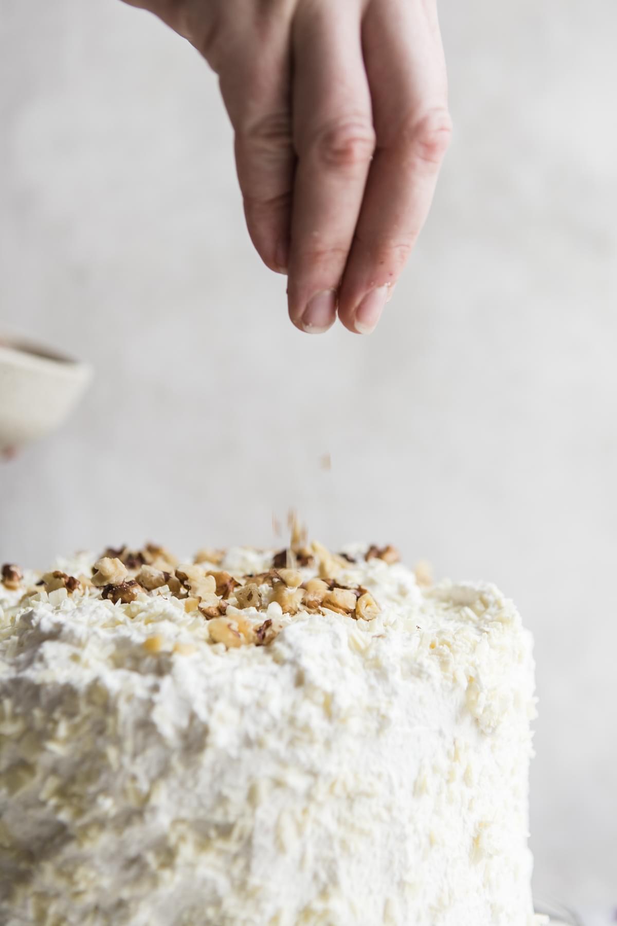 hand sprinkling walnuts over a homemade banana cake with whip cream frosting