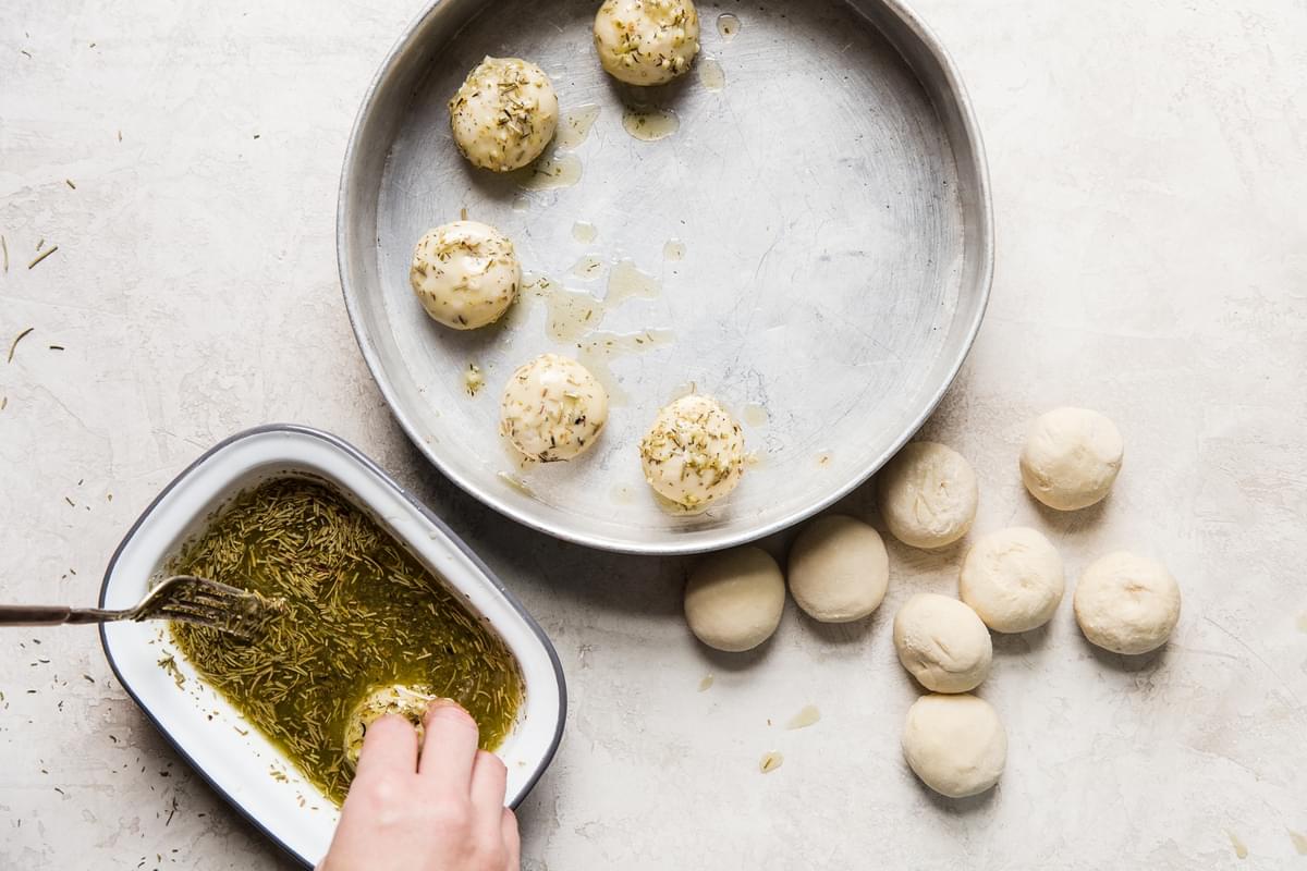 hand rolling frozen dinner rolls in a herb and garlic melted butter and placing them in a pan.