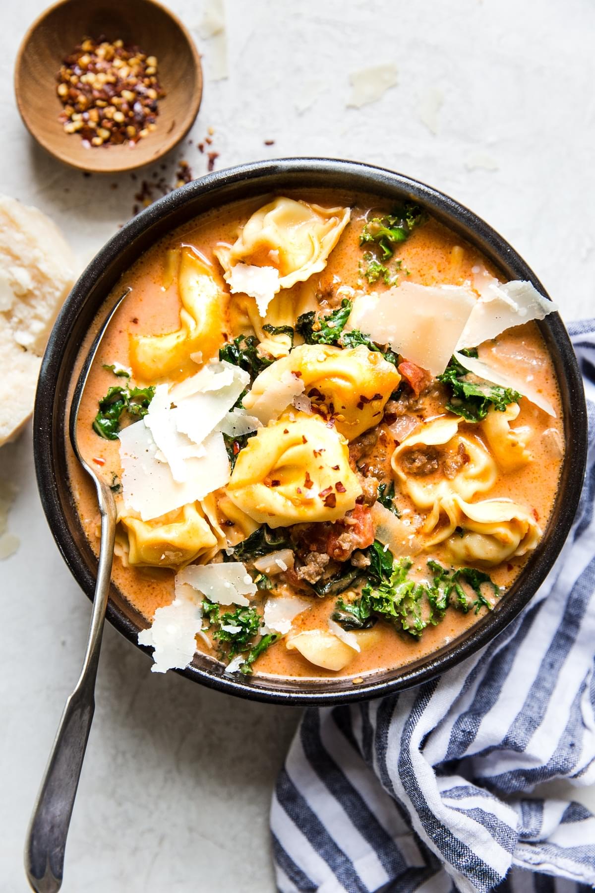 a bowl of tortellini soup with Italian sausage and kale topped with shaved parmesan next to red pepper flakes for serving