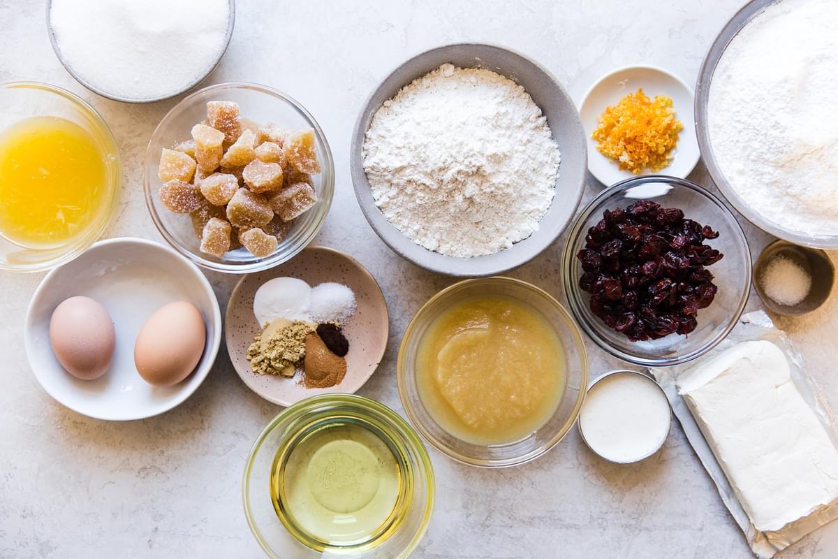 ingredients laid out for gingerbread cake flour, sugar, eggs, oil, apple sauce, cream cheese, orange, cranberries, ginger