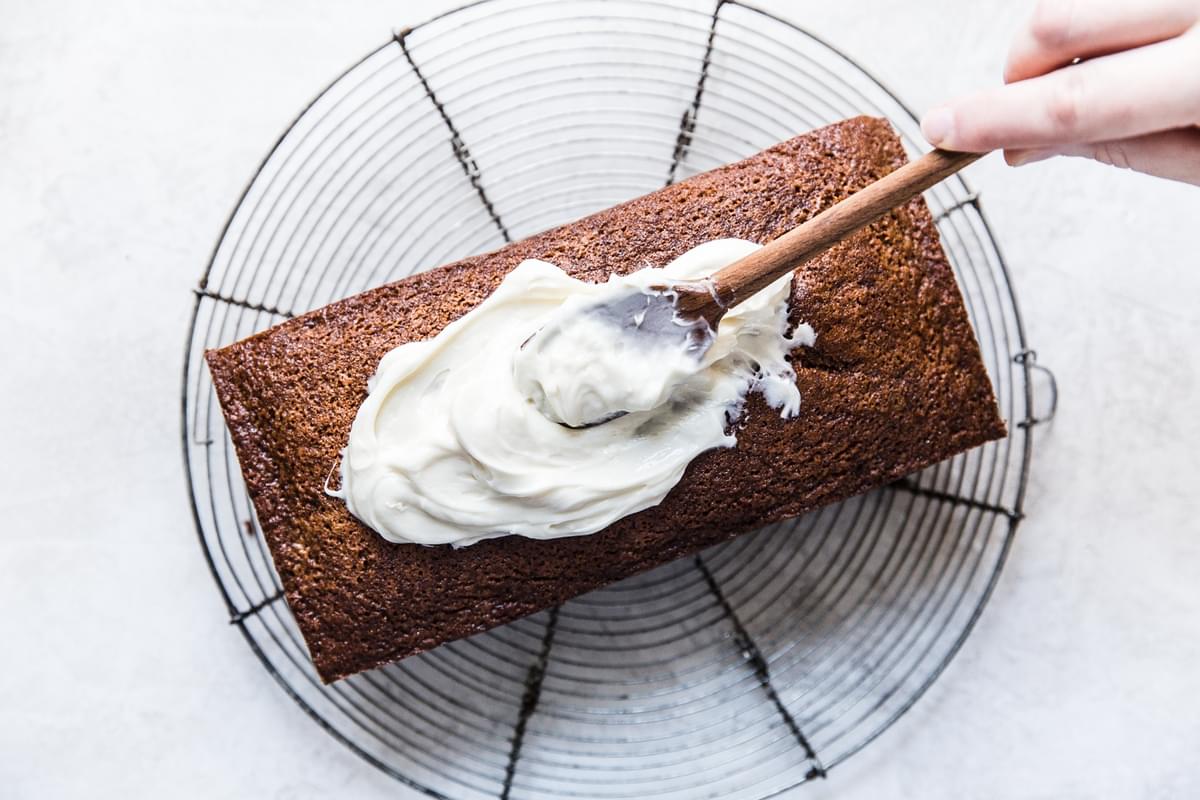 hand spreading cream cheese frosting onto a gingerbread loaf sitting on a cooling rack
