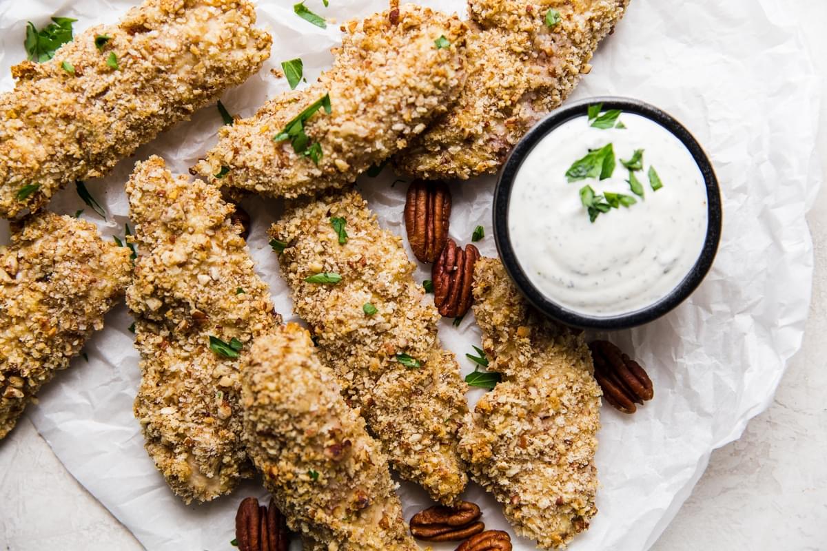 Pecan Crusted Chicken Tenders on parchment paper with a small bowl of buttermilk ranch next to them