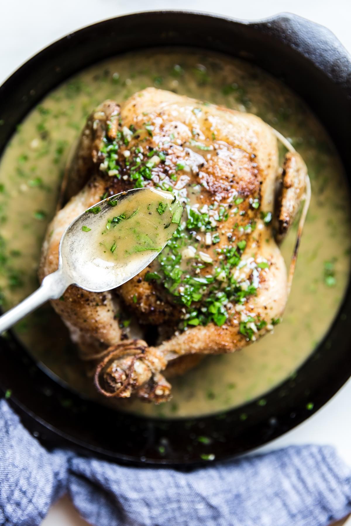 spoon pouring lemon herb pan sauce over a whole roast chicken in a pan.
