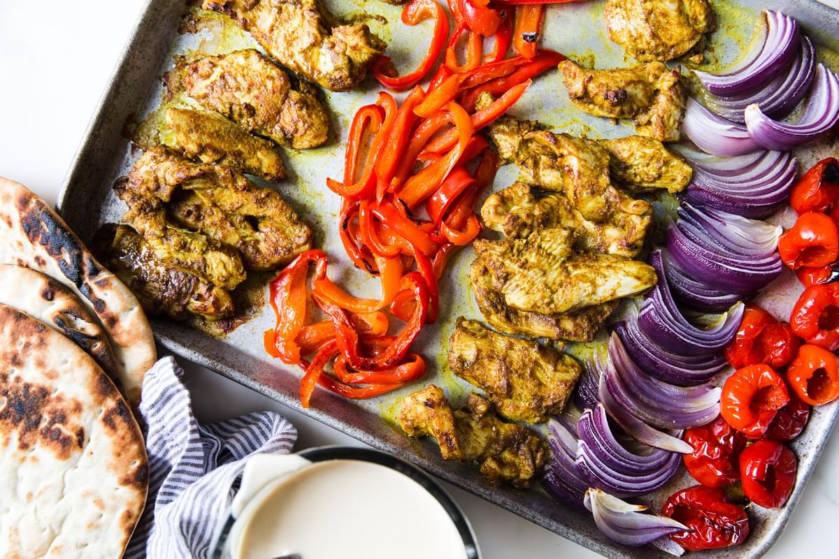 Sheet pan chicken shawarma with red bell peppers and red onion on a baking sheet and tahini sauce in a bowl with pita