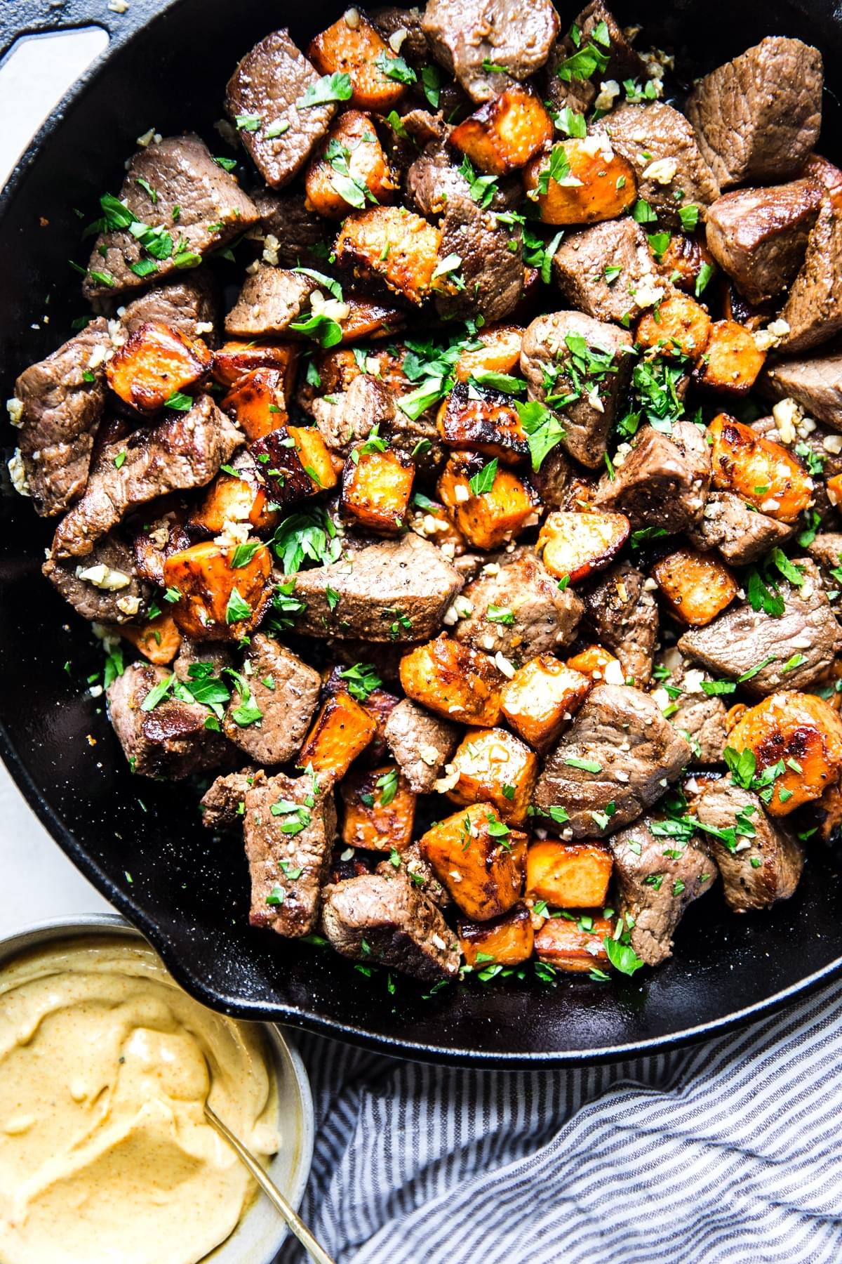 Garlic steak bites with sweet potatoes in a cast iron pan next to curry aioli in a small bowl.