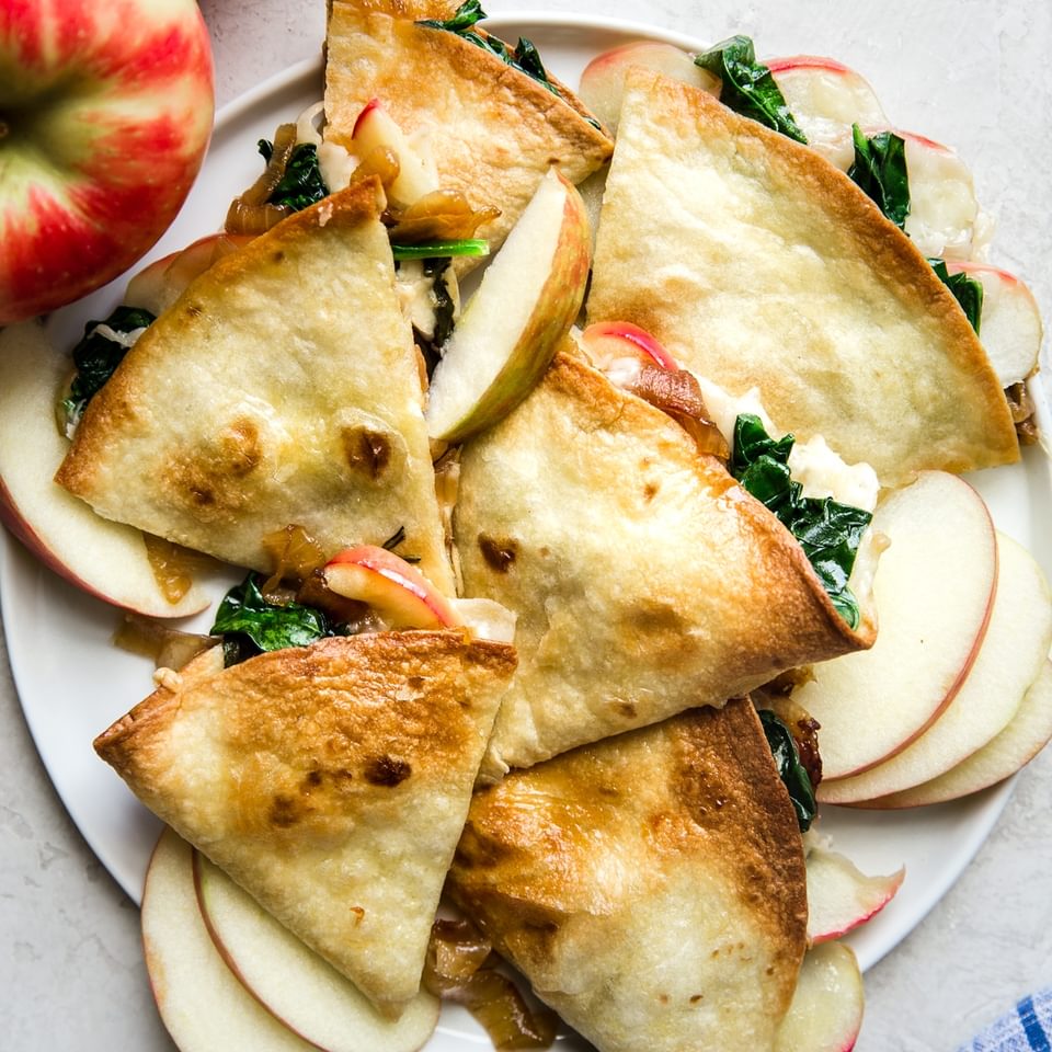Apple, spinach, quesadilla with caramelized onions on a white plate next to apples and a blue linen