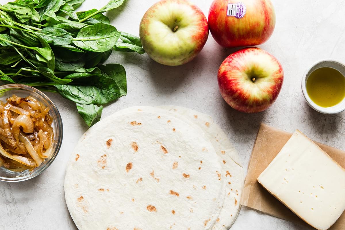Ingredients laid out apples spinach caramelized onions fontina cheese olive oil and tortillas