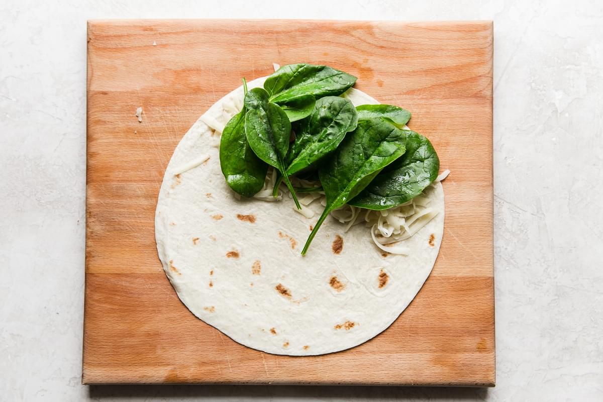 Spinach and fontina cheese on a tortilla on a cutting board.