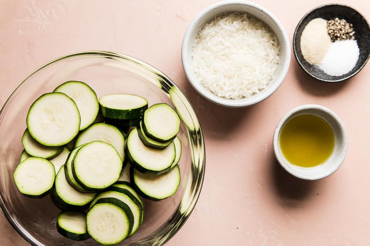 bowl of zucchini slices, bowl of parmesan cheese, bowl with salt pepper and garlic powder and a bowl of olive oil