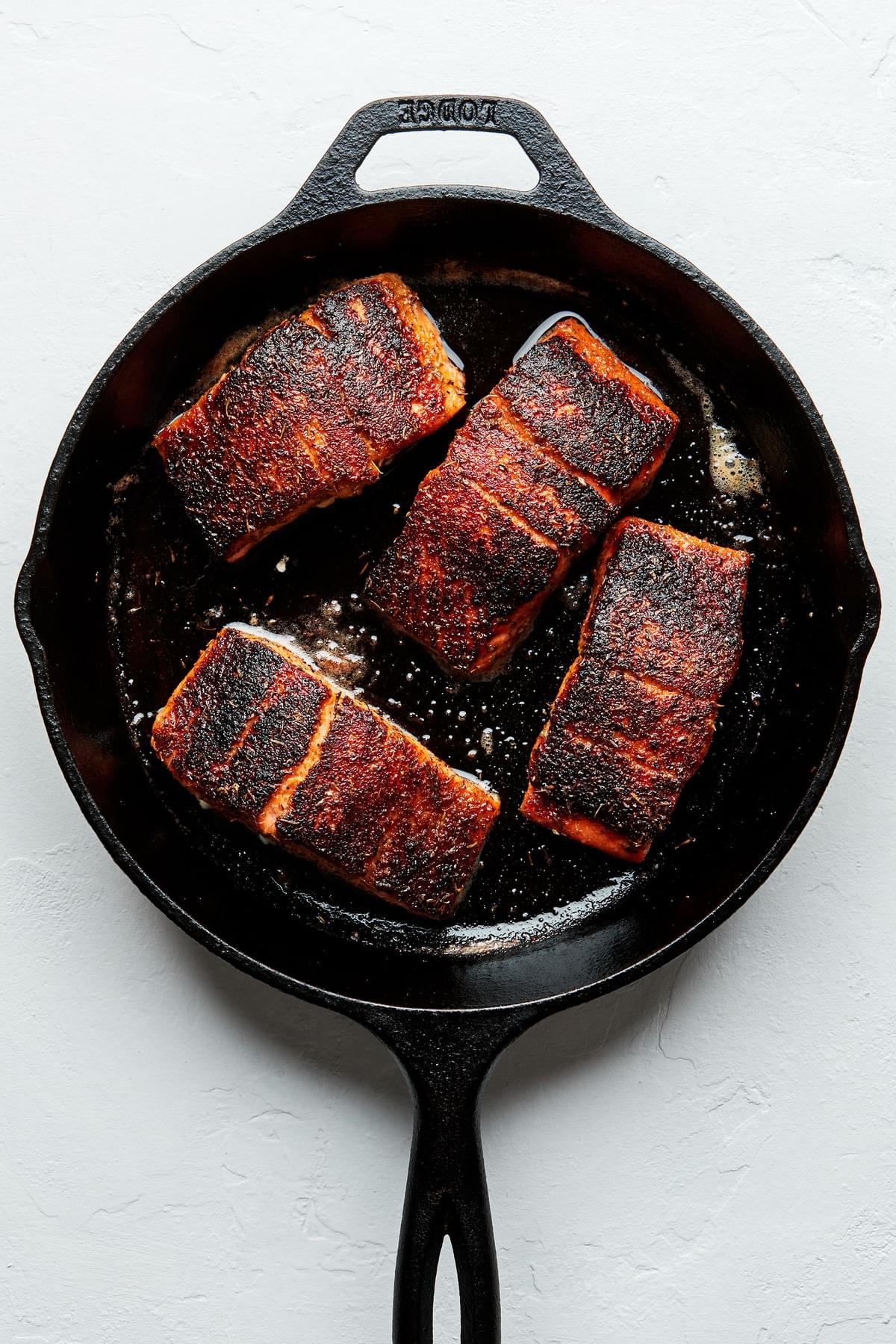 4 blackened salmon filets being cooked in olive oil and melted butter in a cast iron skillet