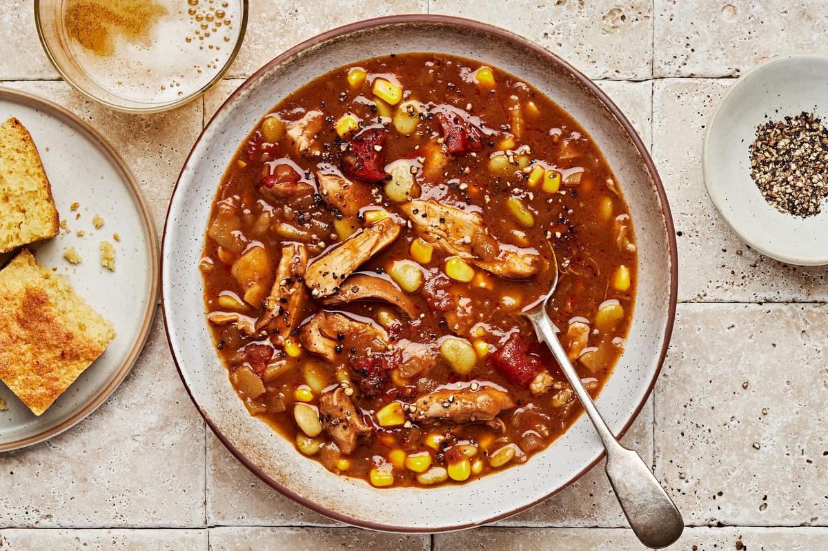 a bowl of homemade Brunswick stew with a spoon next to a small bowl of cracked pepper for serving and a plate of cornbread