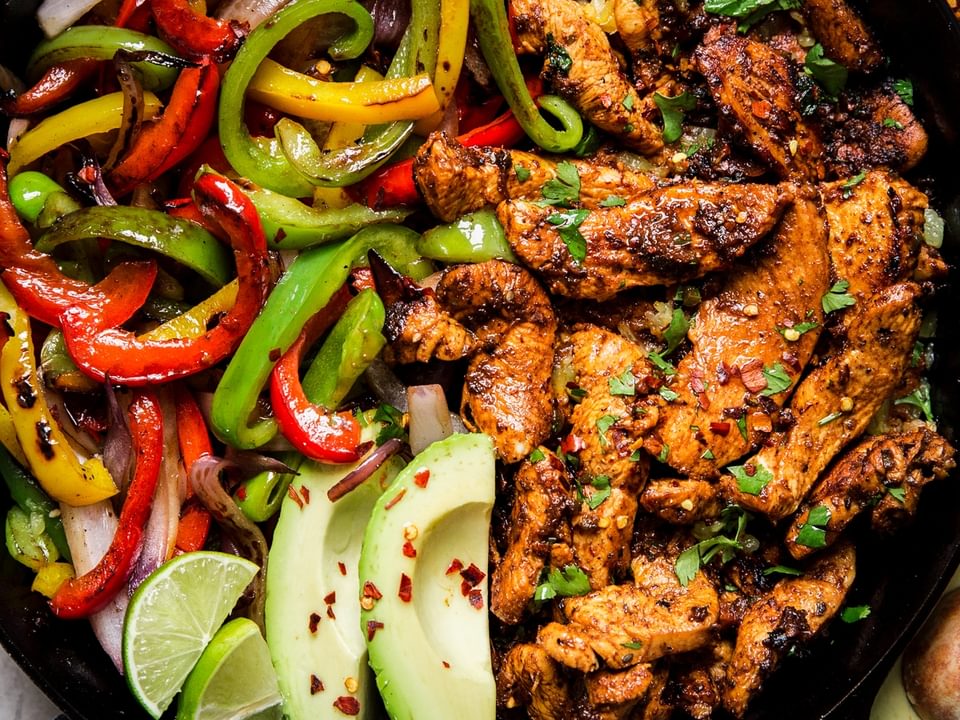 cast iron skillet of chicken fajitas with bell peppers, avocado, and fresh limes