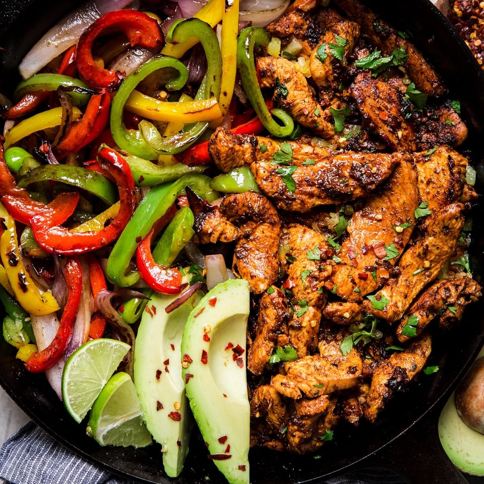 cast iron skillet of chicken fajitas with bell peppers, avocado, and fresh limes