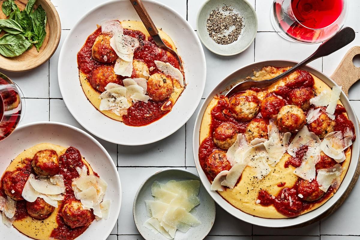 A serving bowl full of chicken meatballs with polenta and marinara being dished up into bowls next to a bowl of parmesan