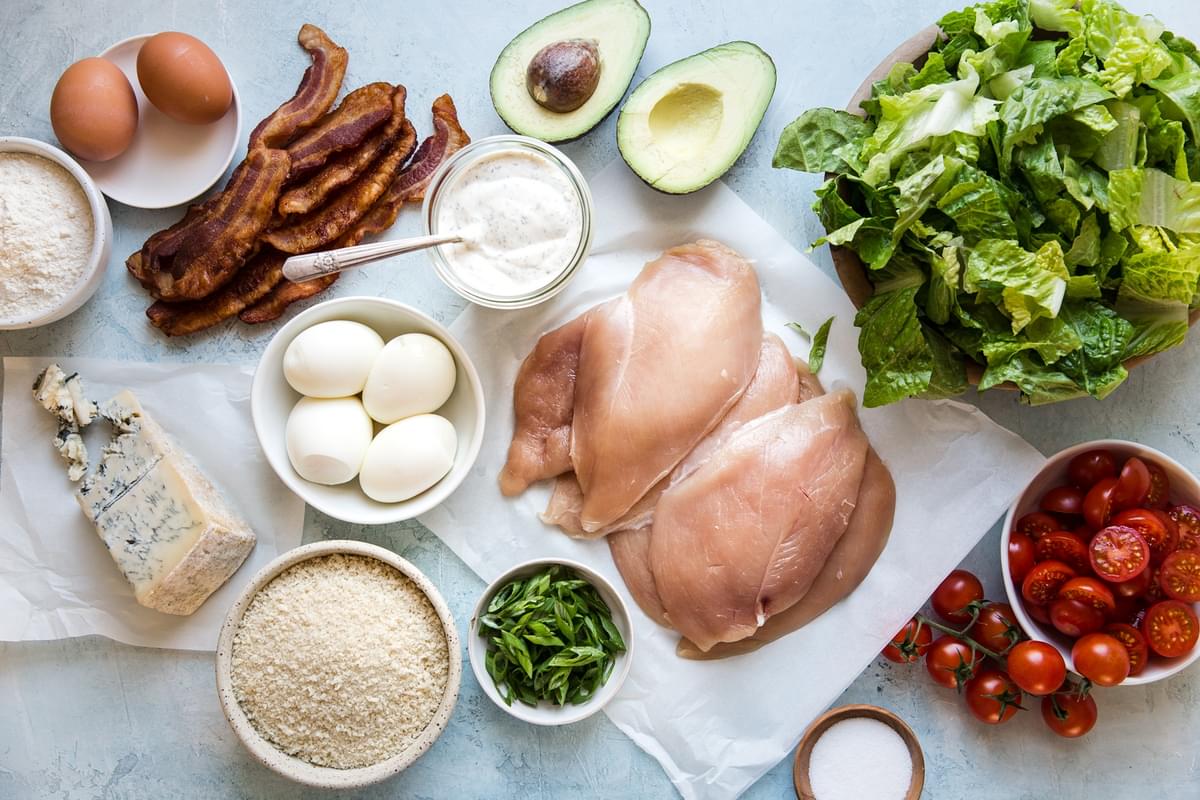 ingredients for classic cobb salad laid out on a table, chicken, romaine, hard boiled eggs, blue cheese, bacon and avocado