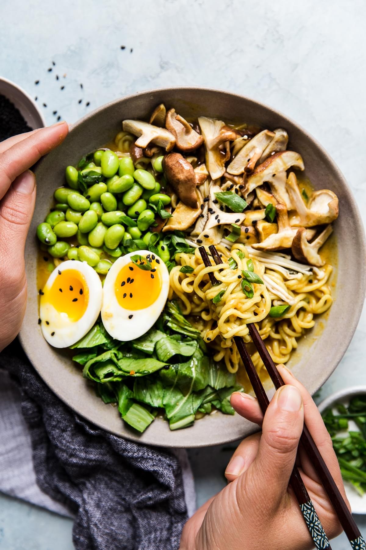 hands using chopsticks to eat Bowl of vegetarian coconut curry ramen with mushrooms, bok choy, edamame and soft boiled egg