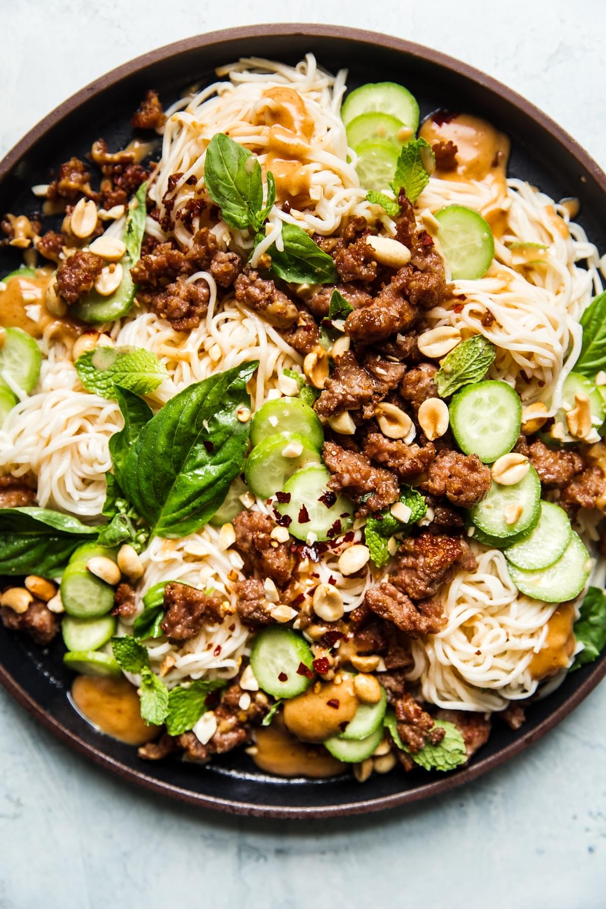 Black plate of Cold Thai noodle salad topped with peanut dressing, crushed peanuts, ground pork, cucumbers, basil and mint