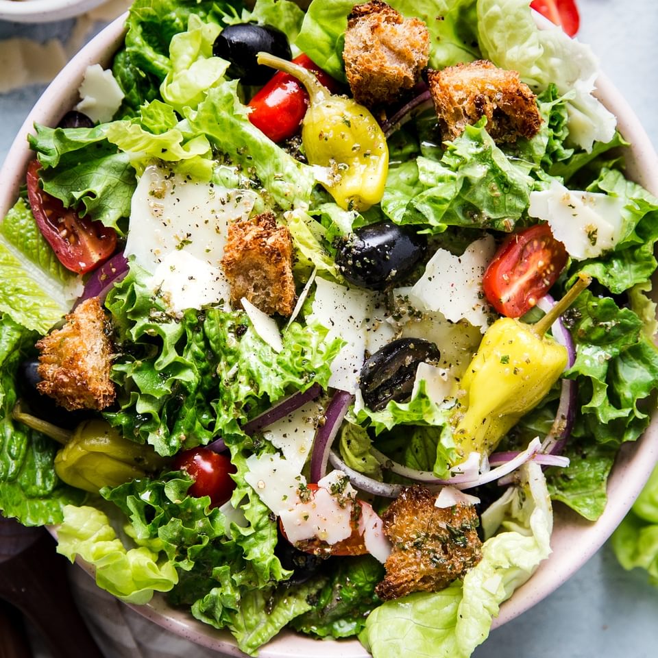 Italian Salad with romaine lettuce, croutons, tomatoes, pepperoncini, black olives, parmesan cheese and red onions in a bowl.