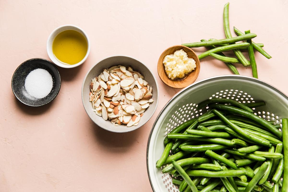 A colander filled with fresh green beans next to minced garlic, sliced almonds, salt and olive oil in small bowls