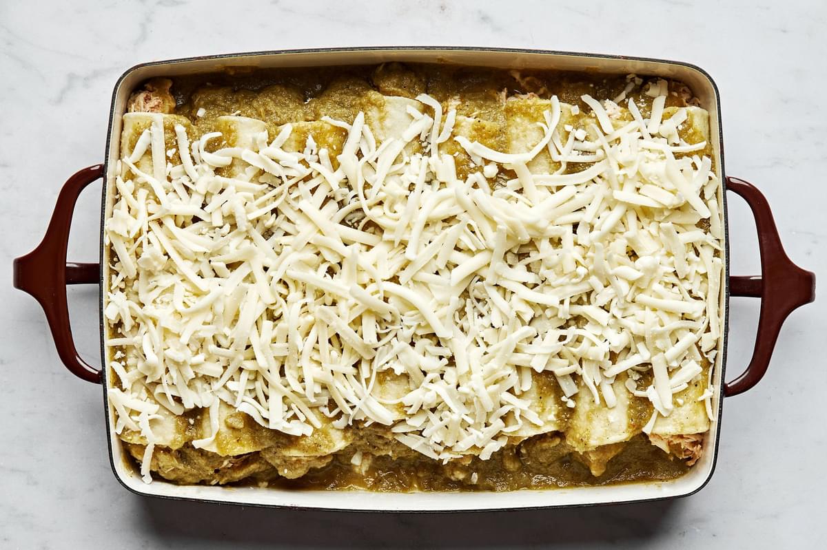rolled green chicken enchiladas lined up in a casserole dish topped with green enchilada sauce and shredded cheese