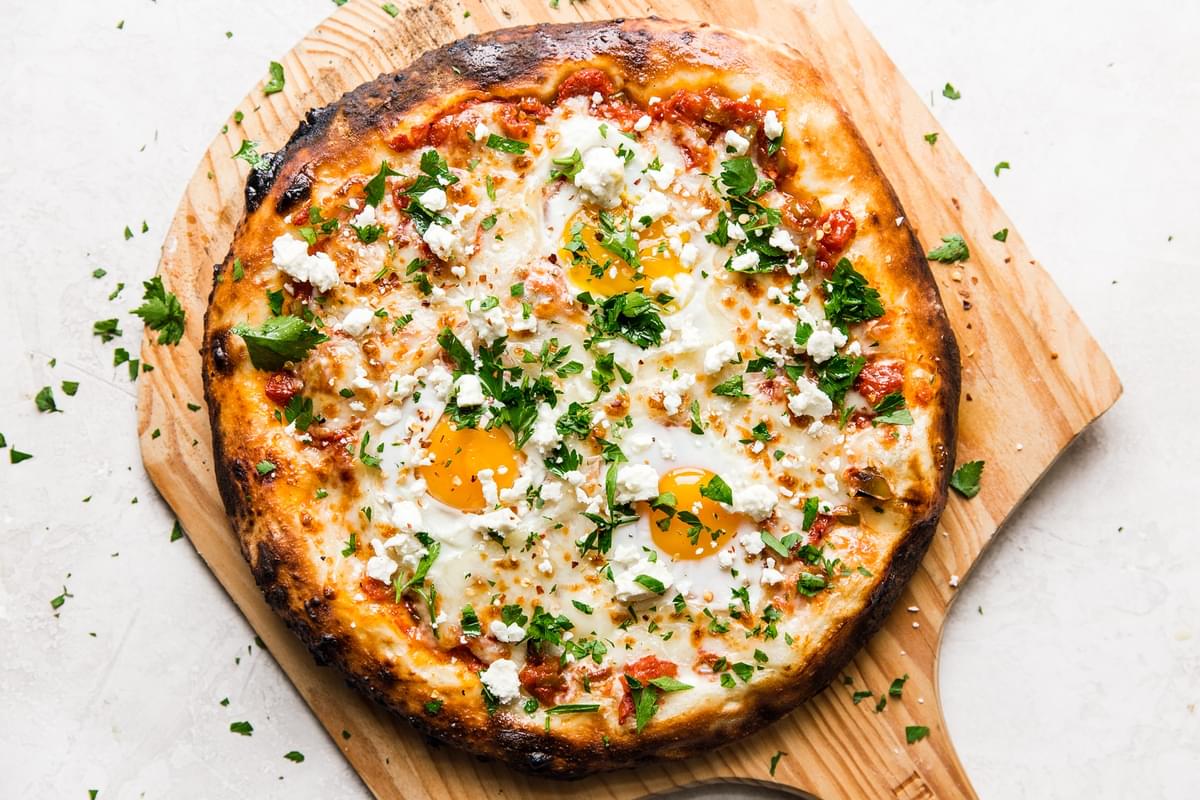 grilled shakshuka pizza on a wood pizza peel topped with fresh parsley and feta