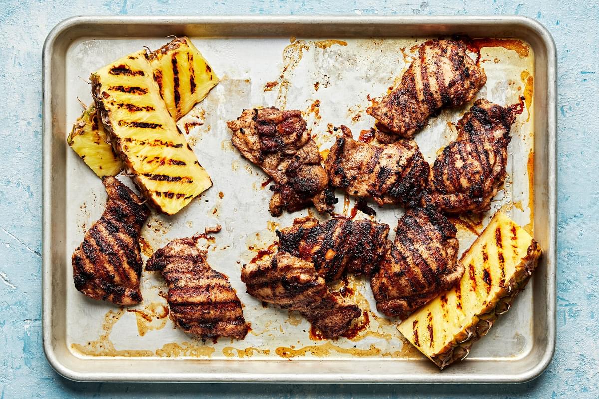 grilled jerk chicken thighs and grilled pineapples wedges on a baking sheet