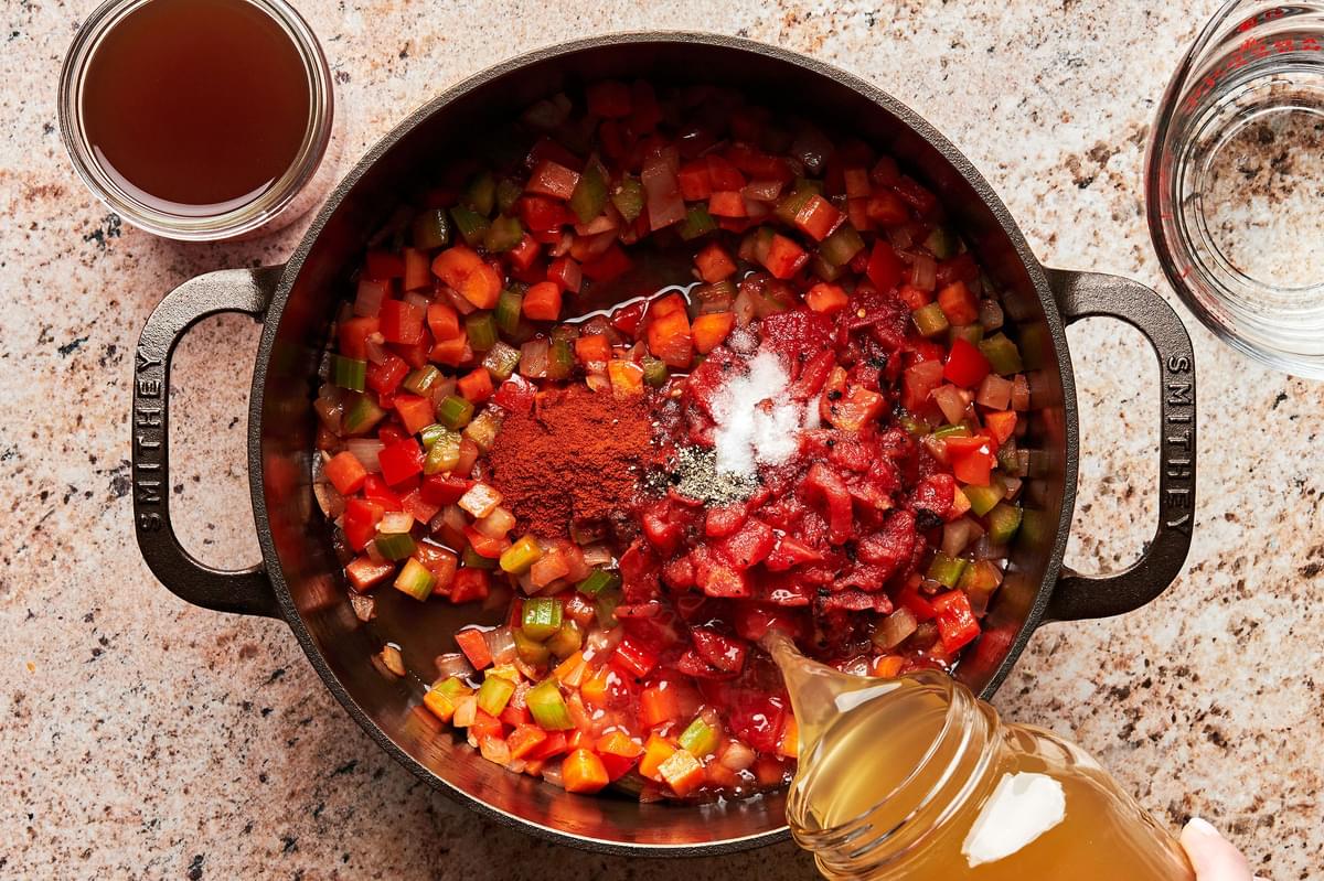 stock being poured into a pot with onion, celery, carrots, bell pepper, tomato paste, tomatoes, paprika, salt and pepper