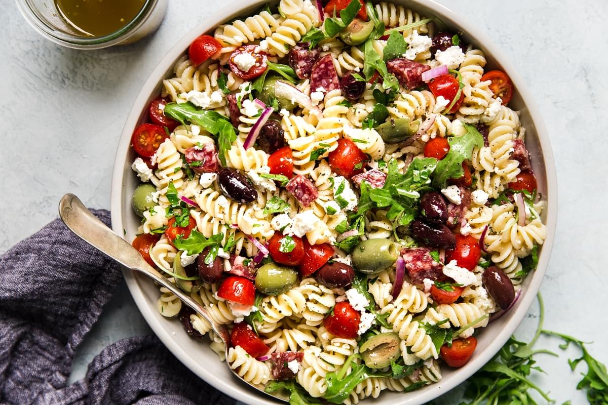 Mediterranean Pasta Salad in a bowl with feta, olives, tomatoes and arugula with a serving spoon