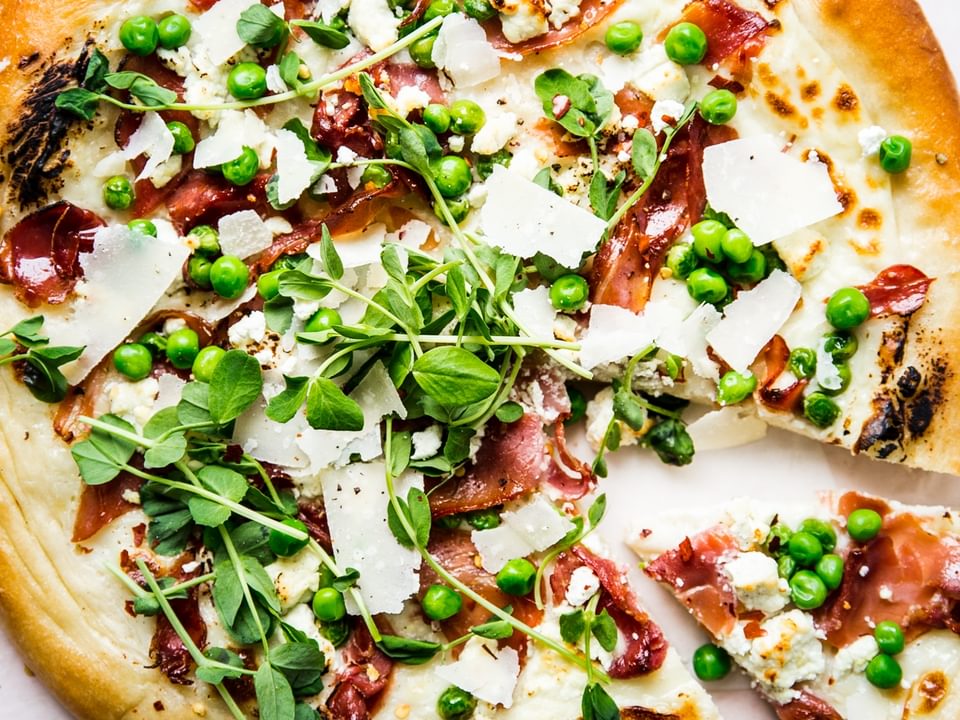 Pea and prosciutto pizza with goat cheese cream sauce parmesan cheese spring pizza with one slice taken out.