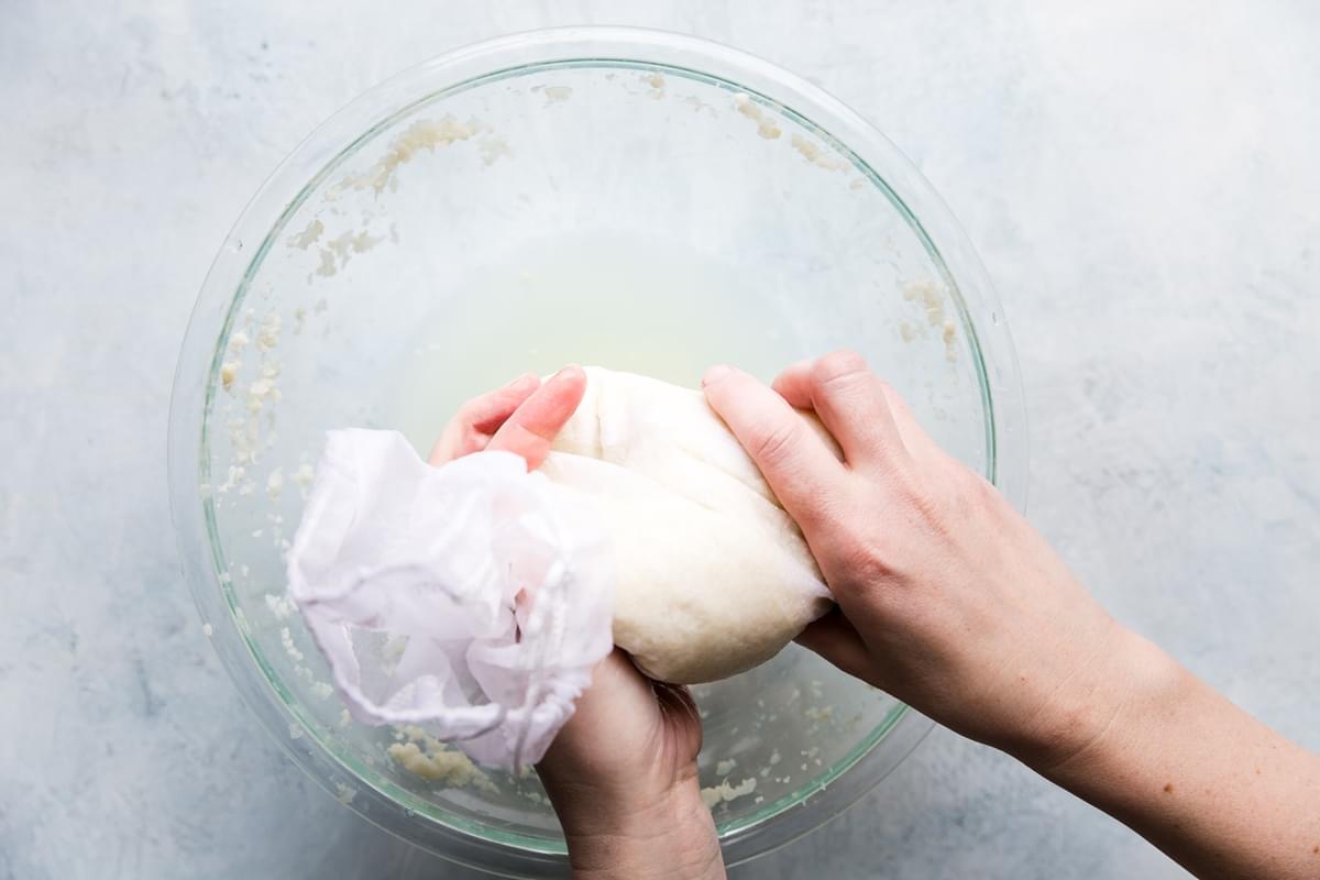 two hands squeezing liquid out of cooked cauliflower into a bowl with a nut bag