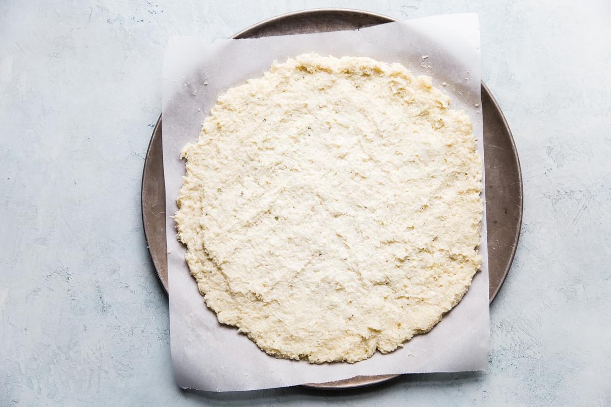 raw cauliflower pizza crust on a piece of parchment on a pizza pan
