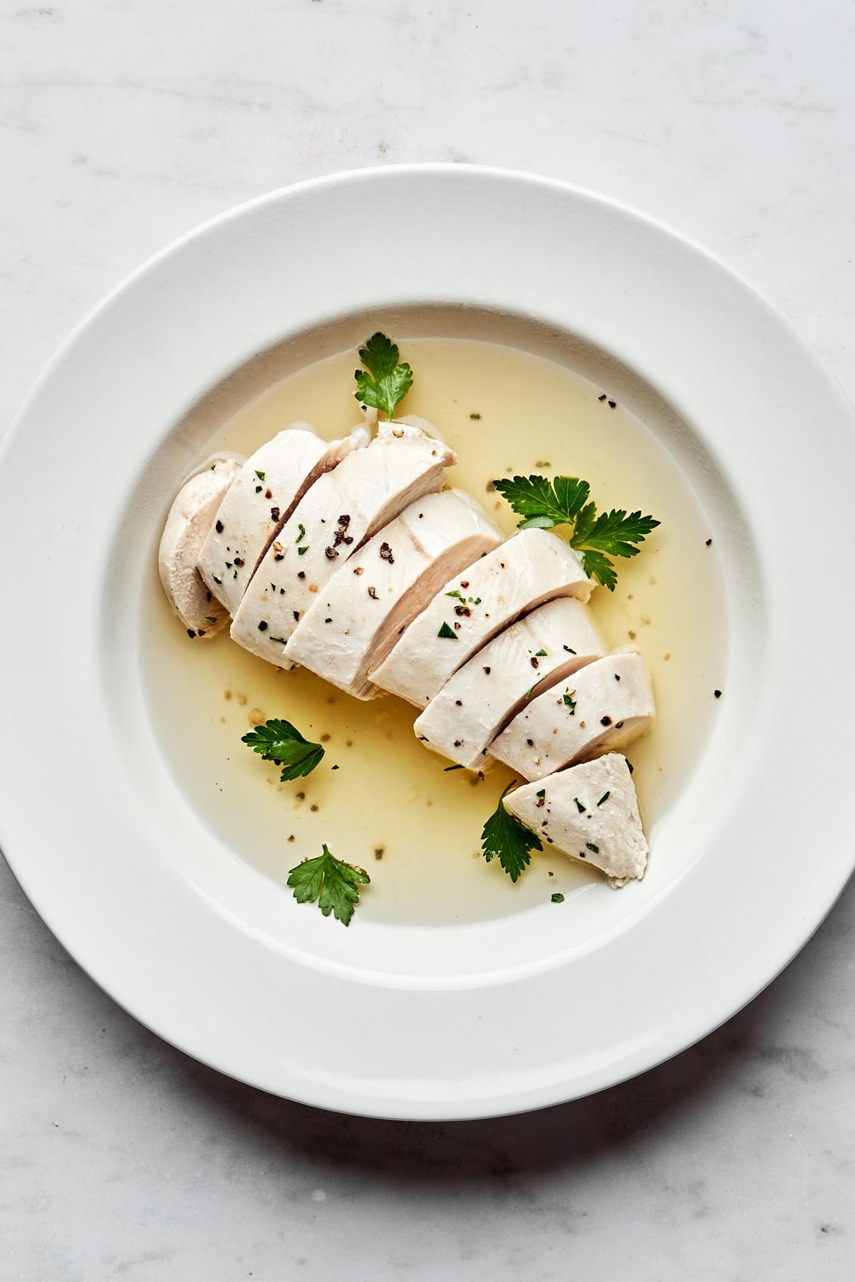 perfect poached chicken sliced in a bowl with poaching liquid cooked in chicken broth, salt, bay leaf, peppercorns and garlic