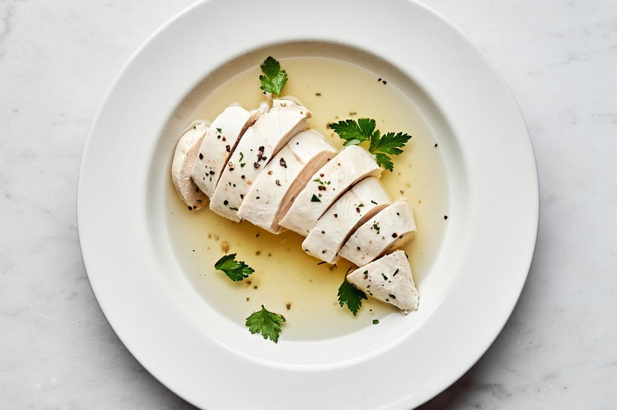perfect poached chicken sliced in a bowl with poaching liquid cooked in chicken broth, salt, bay leaf, peppercorns and garlic