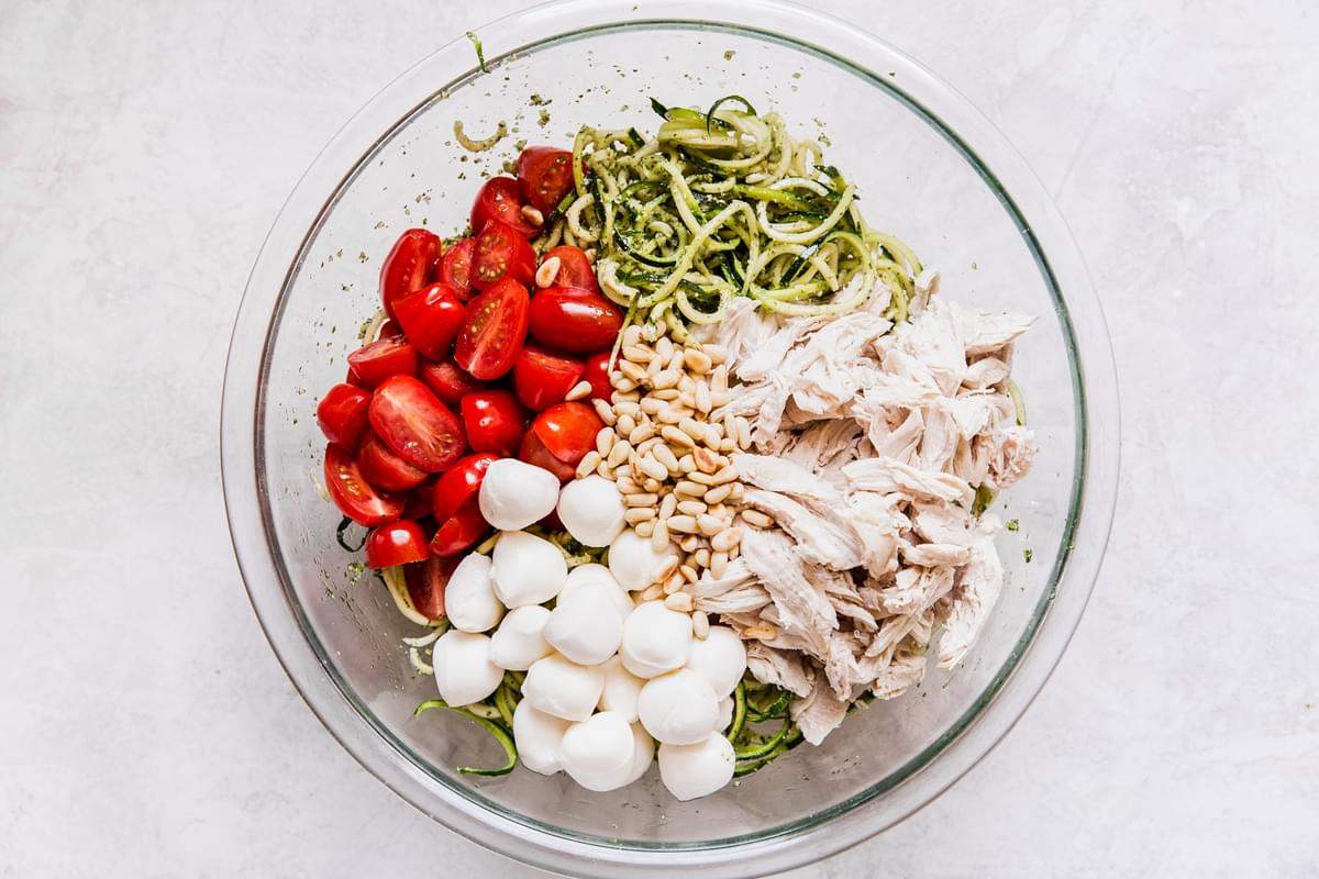 glass bowl filled with zoodles, pesto, shredded chicken, pine nuts, cherry tomatoes and pearl mozzarella