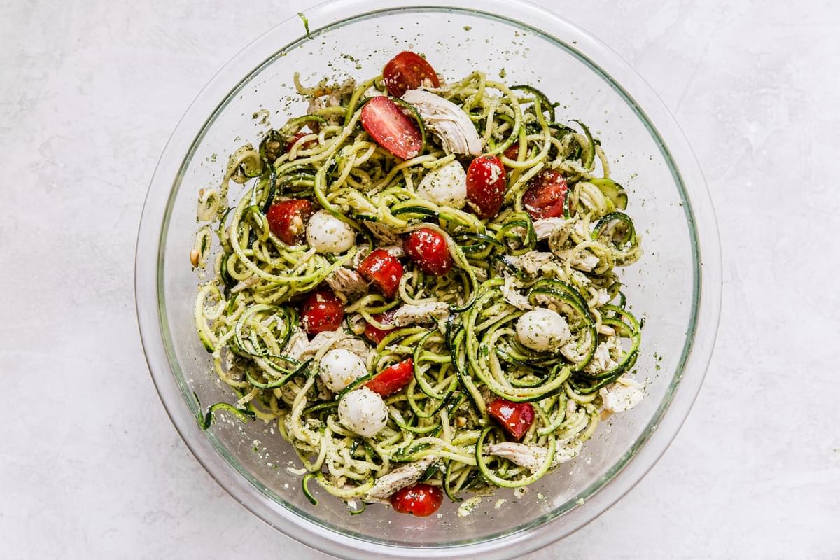 glass bowl filled with Pesto chicken zoodle salad topped with fresh cherry tomatoes, pearl mozzarella and toasted pine nuts