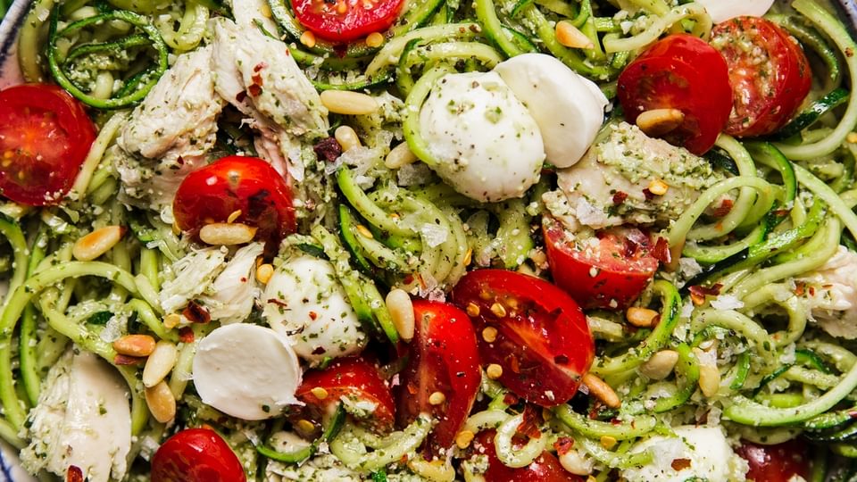 white bowl filled with Pesto chicken zoodle salad topped with fresh cherry tomatoes, pearl mozzarella and toasted pine nuts