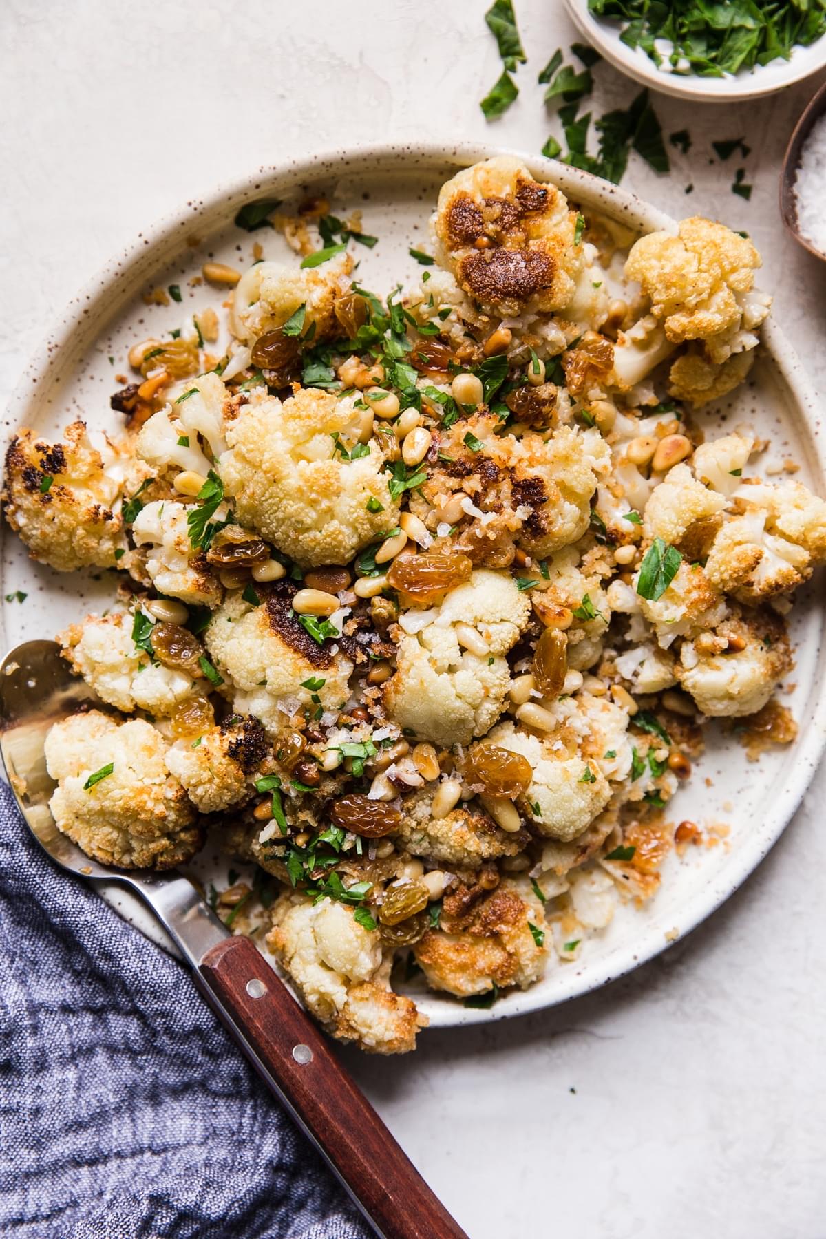 Roasted Cauliflower With Panko And Golden Raisins on a plate with a fork next to a bowl of fresh minced parley