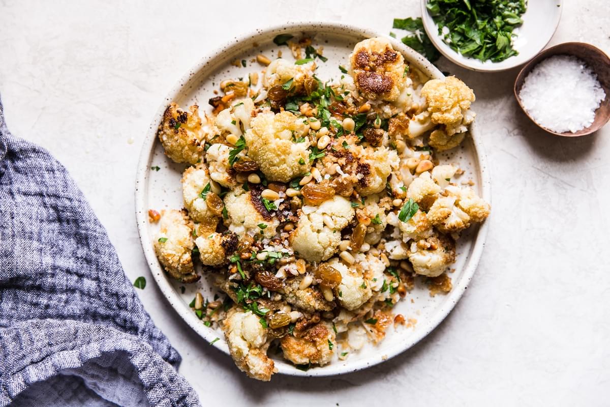 Roasted Cauliflower With Panko And Golden Raisins on a plate with a linen next to a bowl of parsley and a bowl of salt