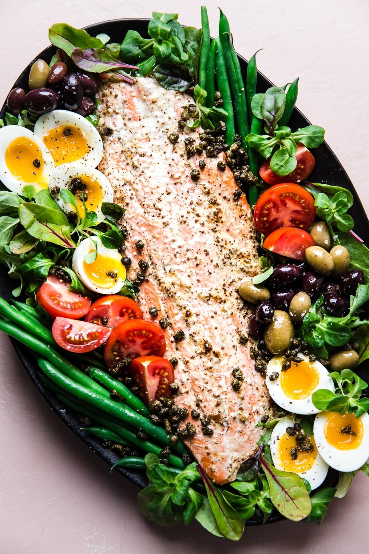 Salmon Niçoise Salad on a platter with olives, tomatoes, green beans, fried capers and soft boiled eggs.