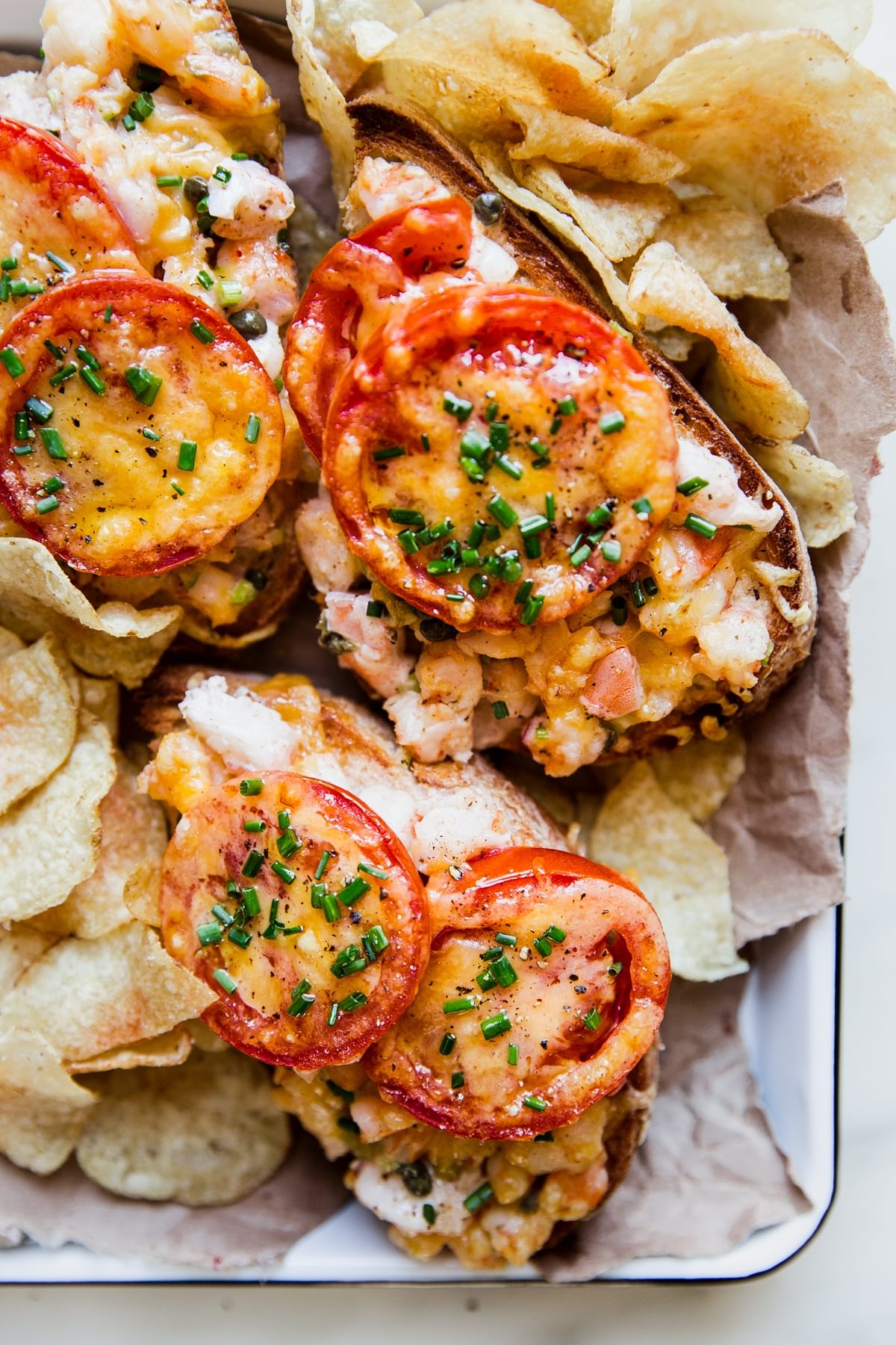 three open faced shrimp melts topped with fresh tomato slices and fresh chives on a tray surrounded by potato chips