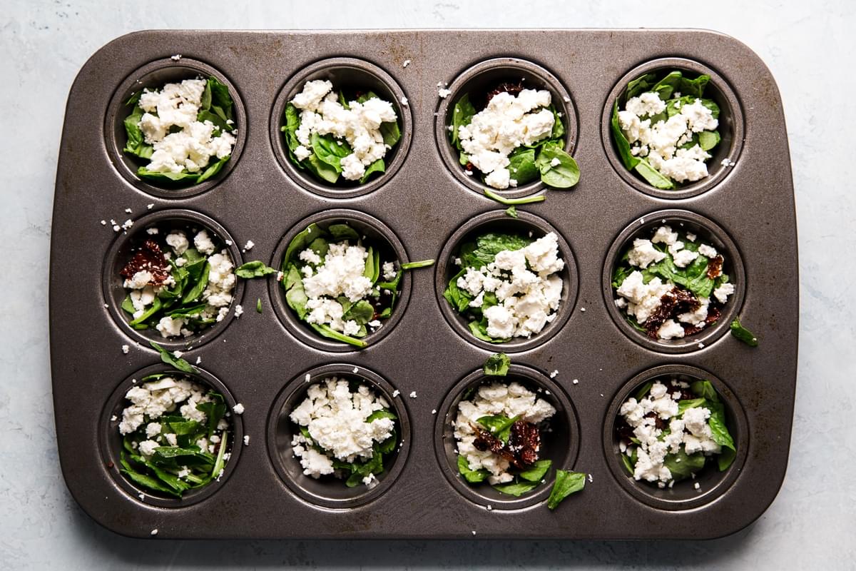 feta, spinach and sun-dried tomatoes in muffin tins ready for egg to be added to make breakfast egg cups