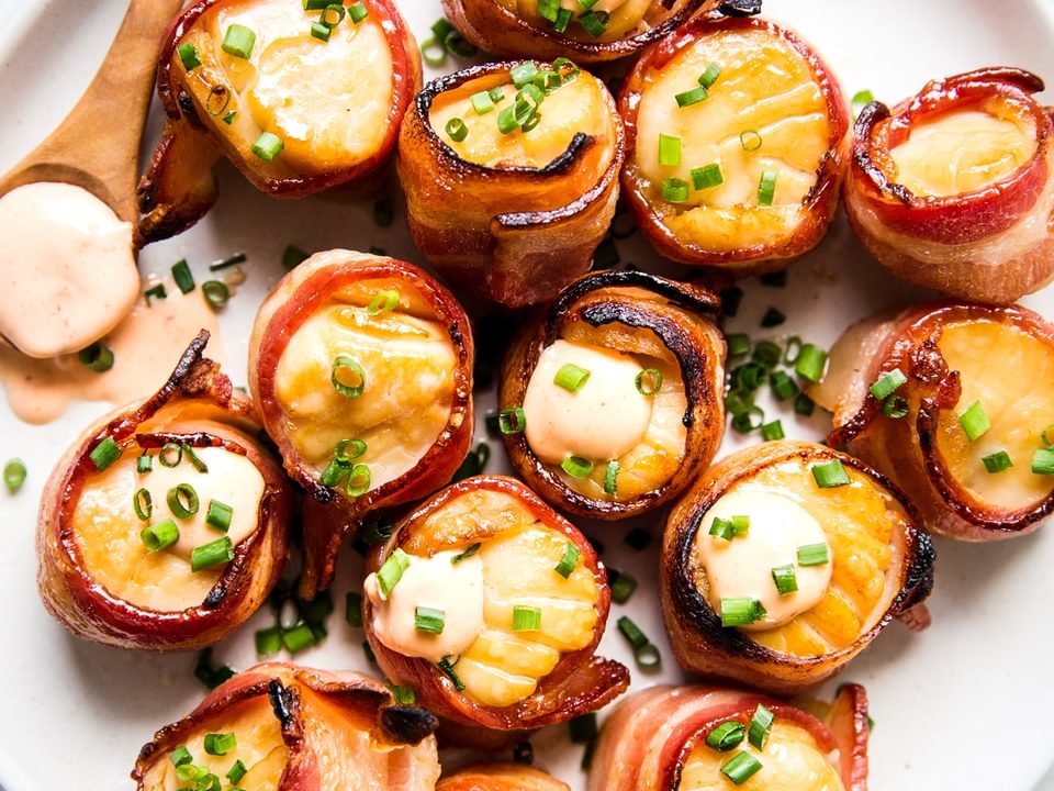 bacon wrapped sea scallops on a white plate with sriracha aioli and topped with fresh chives