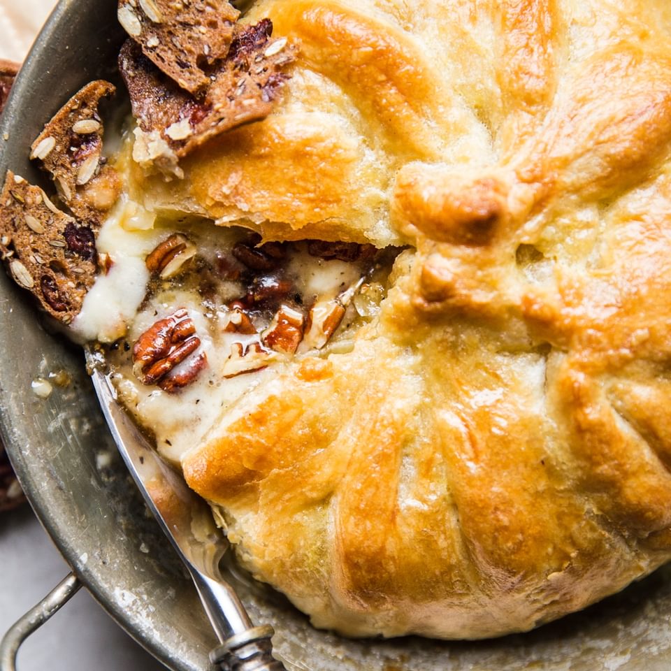 baked brie made with pecans, brown sugar and pepper wrapped in puffed pastry dough in baking dish being served with crackers