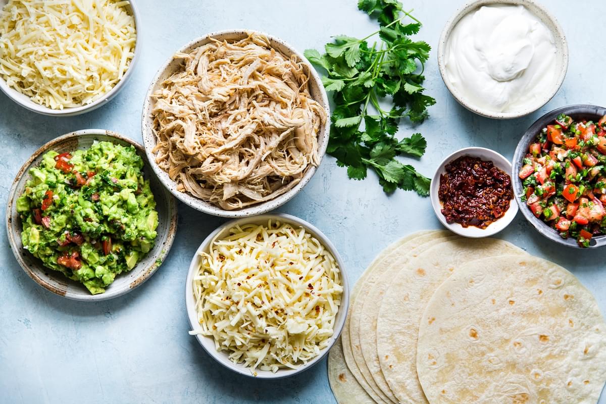 ingredients for chicken quesadillas laid out; guacamole, shredded chicken, flour tortillas, cheese, sour cream, chipotle