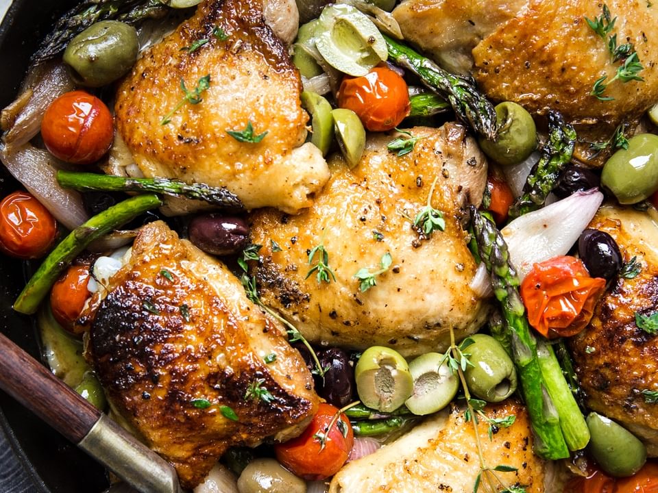 Baked chicken thighs in a cast iron skillet surrounded by asparagus, olives, and tomatoes.