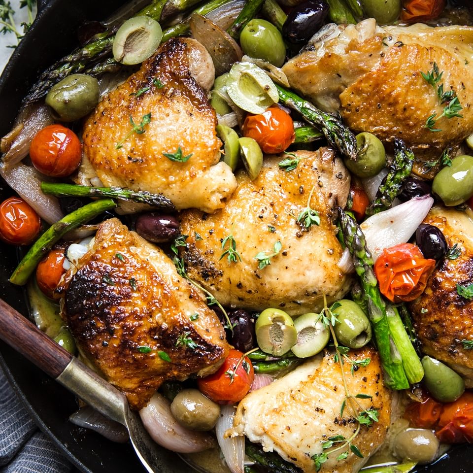 Baked chicken thighs in a cast iron skillet surrounded by asparagus, olives, and tomatoes.