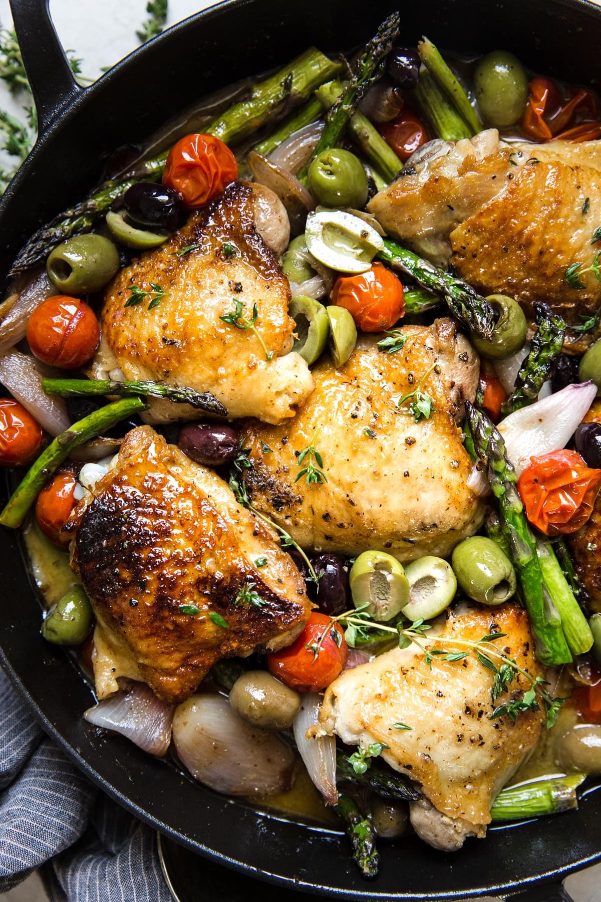 Baked Chicken Thighs With Asparagus in a pan with olives, tomatoes and shallots in a white wine garlic sauce.