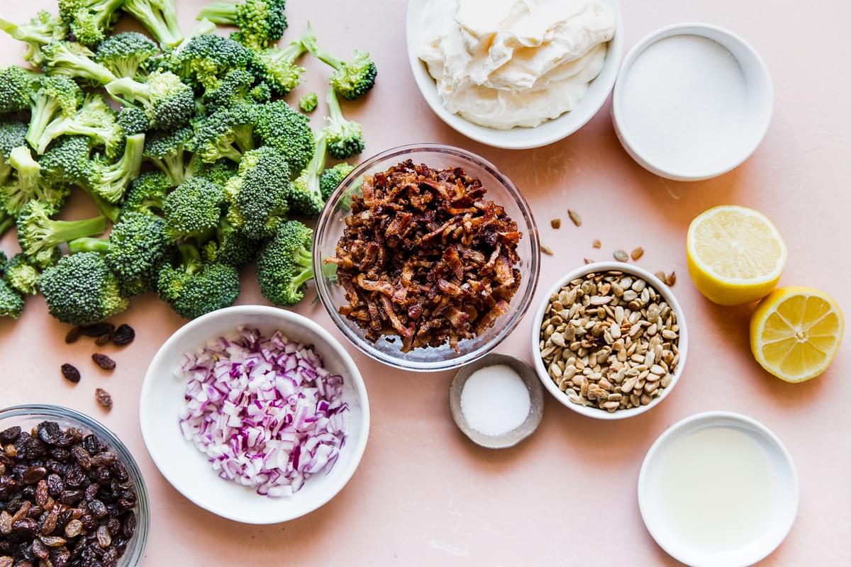 ingredients laid out for broccoli salad; broccoli, bacon, mayonnaise, sugar, lemon juice, onions, golden raisins