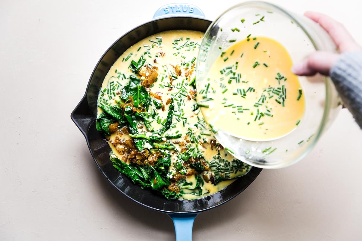 blue cast iron skillet with caramelized onions and baby kale with beaten eggs being poured over the top