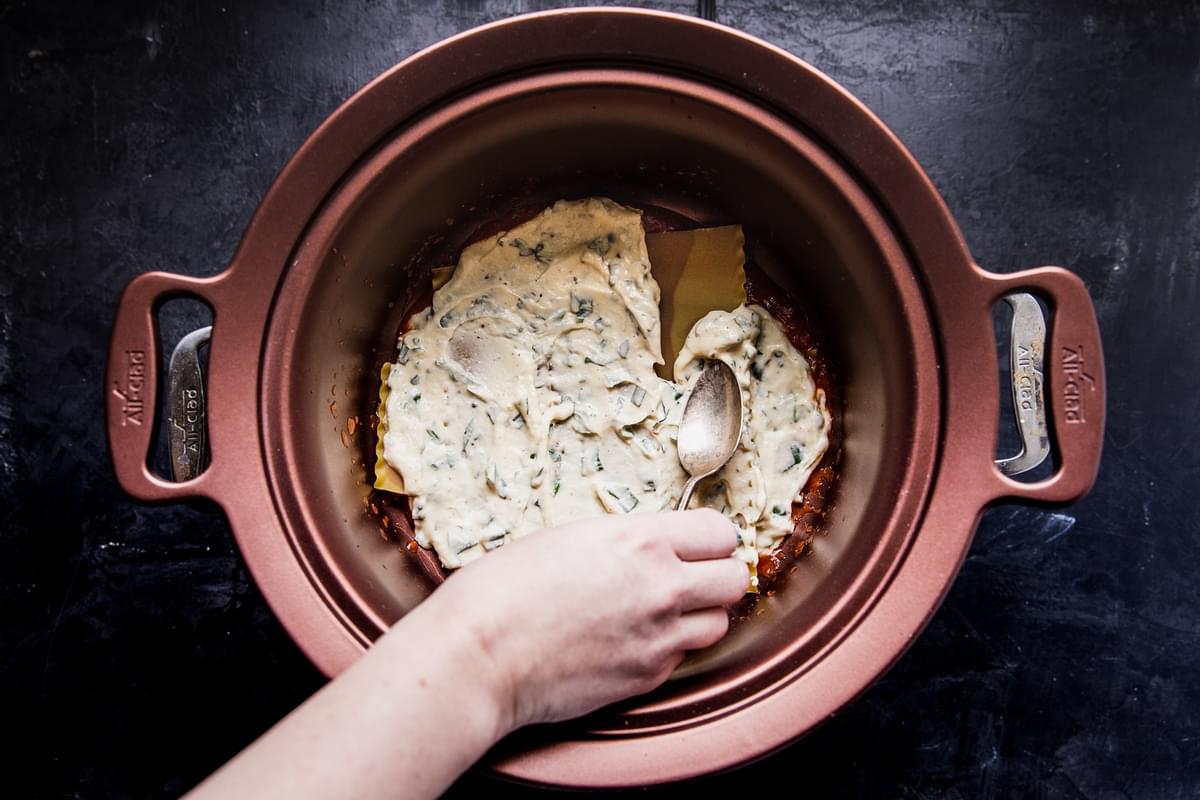 hand spreding ricotta filling on lasagna noodles in a crockpot